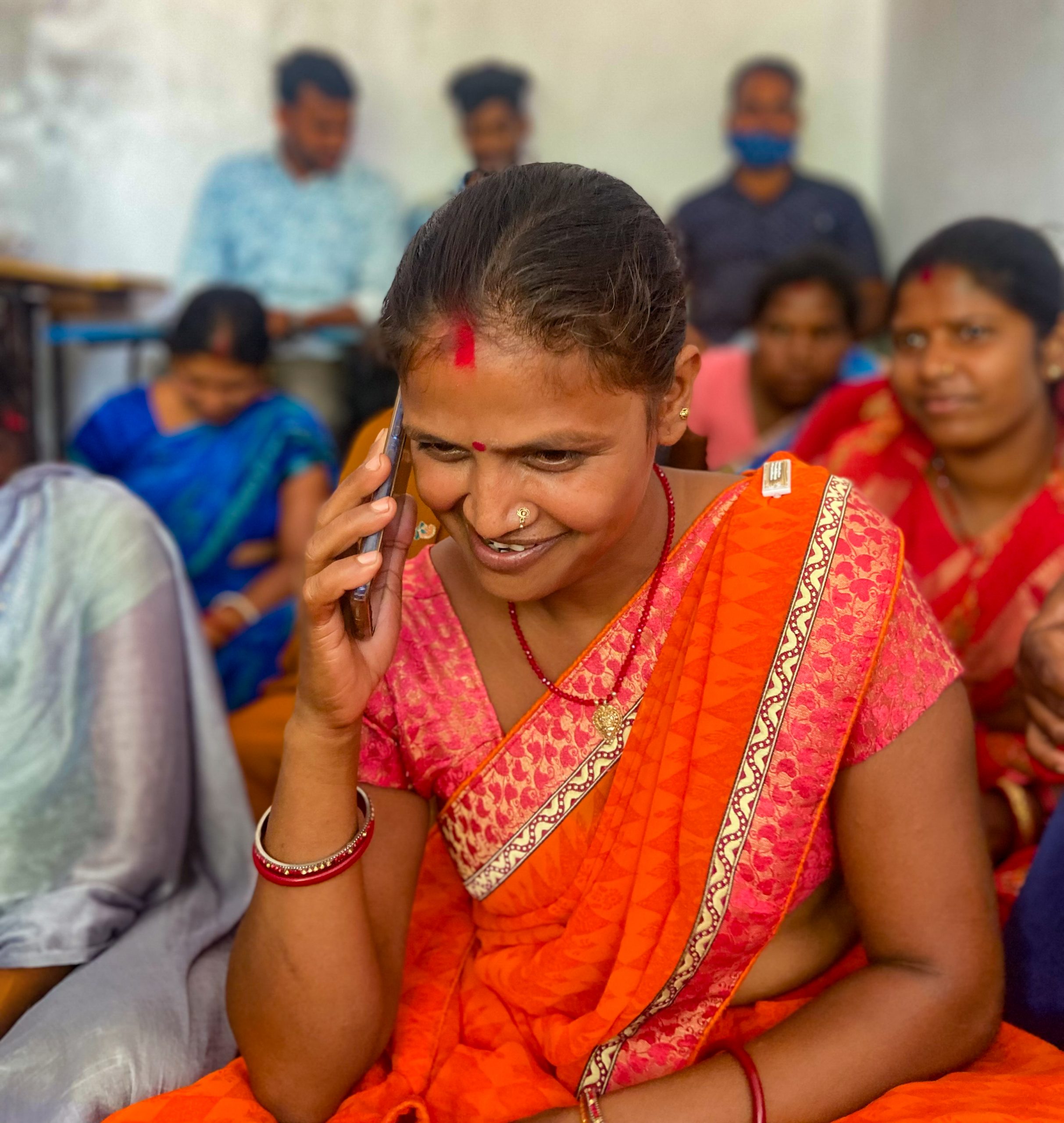 A woman using her phone to call someone while sitting in a room with other people
