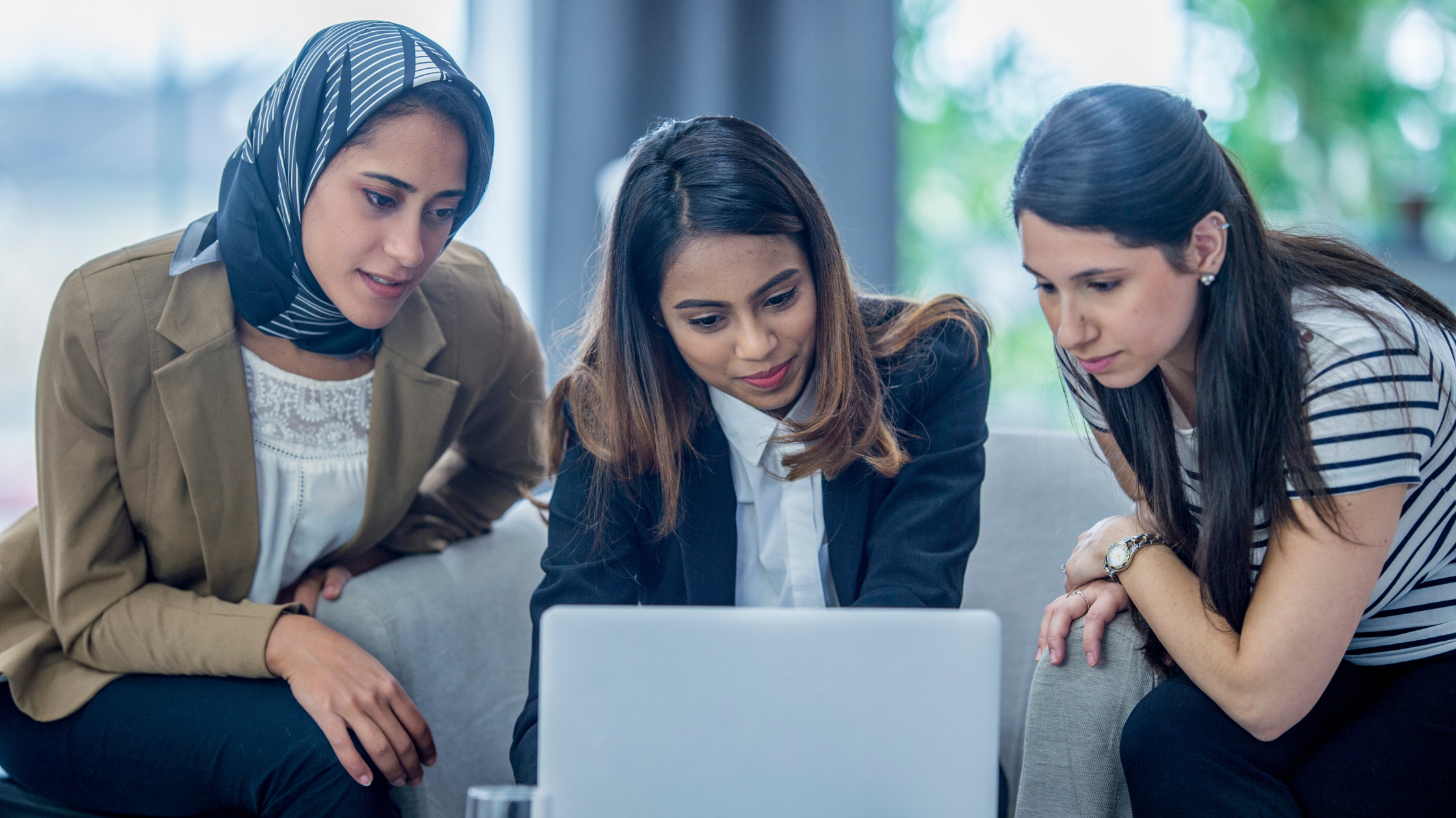 Three women in business attires are indoors in an office work space, sitting around a laptop computer.